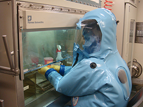 This figure is a photo of a trainee in a bio-containment suit, working in a laboratory.