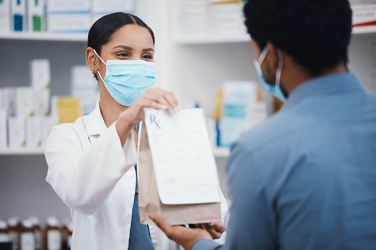 masked pharmacist handing a bagged prescription to a customer
